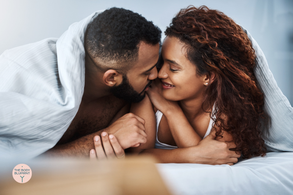 A couple lying in bed, facing each other and smiling, wrapped together in a blanket, expressing affection and intimacy.